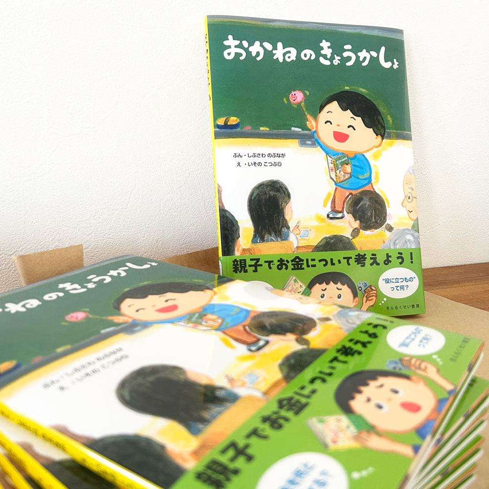 手前にいくつか積み上げてあり、奥に表1側が見えるように1冊立てて置いてある『おかねのきょうかしょ』